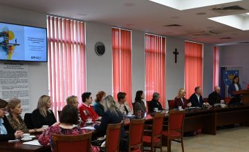Sala konferencyja, przy stole pod oknami siedzą zebrani na konferencji goście